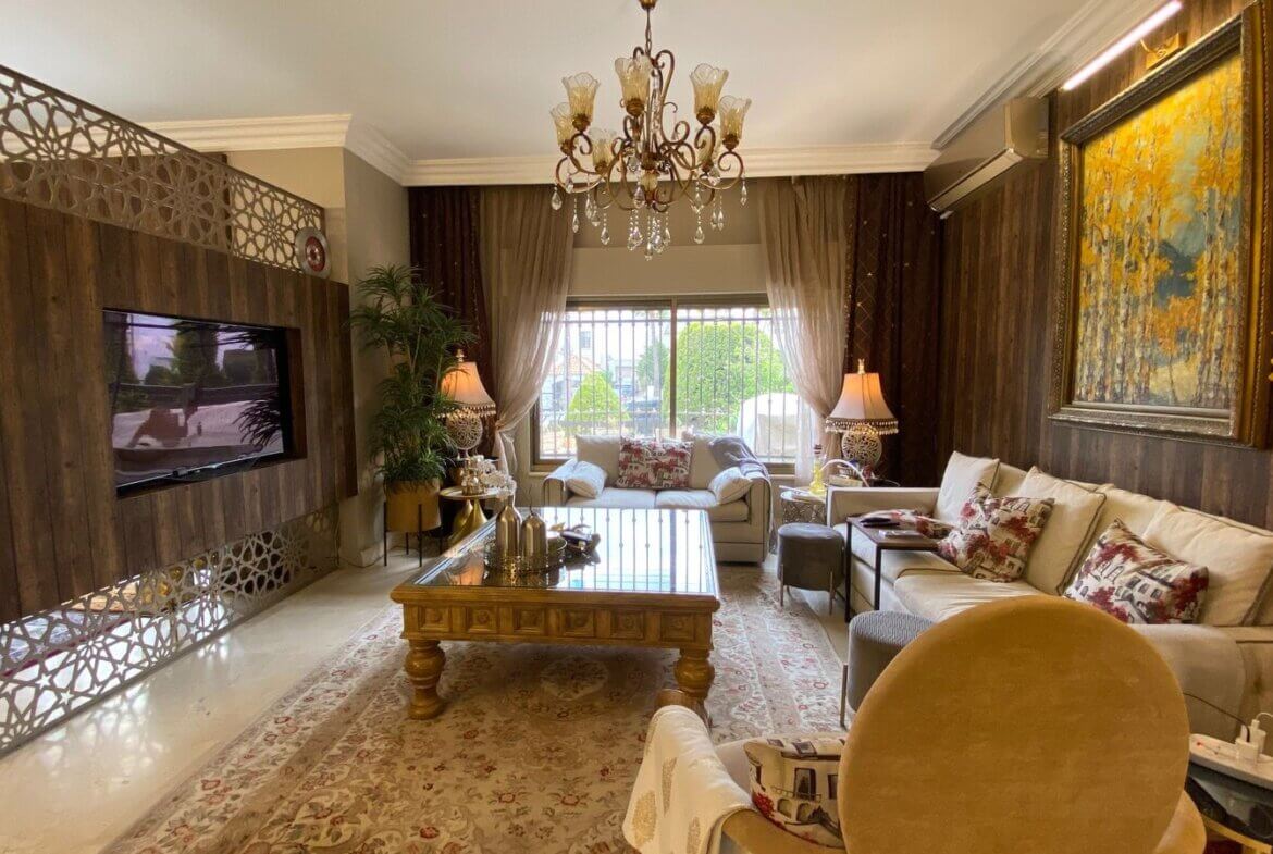 24c9887b-9507-4411-82d8-3200047c2154 Luxurious living room with wood-paneled wall, ornate crystal chandelier, and beige sofas around a glass-topped coffee table.