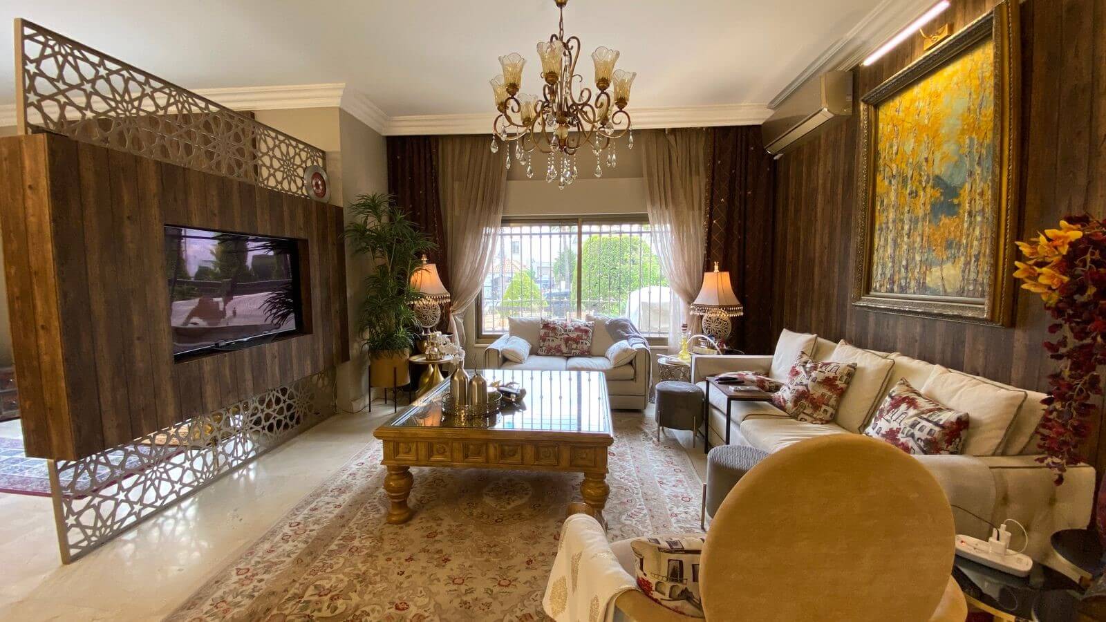 24c9887b-9507-4411-82d8-3200047c2154 Luxurious living room with wood-paneled wall, ornate crystal chandelier, and beige sofas around a glass-topped coffee table.