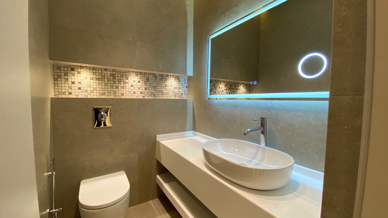 2588ba6a-d65f-4e6e-a707-07126380992a Modern bathroom with a white vessel sink on a floating counter, a wall-mounted toilet, chrome faucet, and a rectangular backlit mirror with blue LED edge lighting; grey tiled walls with a mosaic tile backsplash.