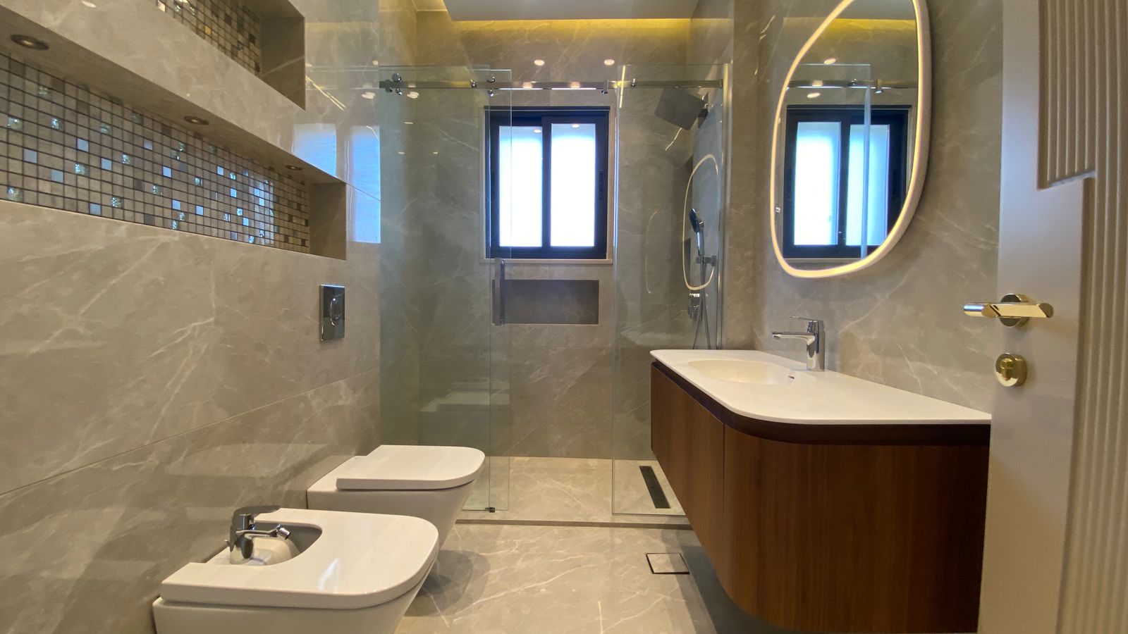 25d24960-2602-46b5-a6fe-f75991b7301a Modern marble bathroom with glass shower enclosure, wall-hung toilet and bidet, and a curved wooden vanity topped with a white sink and an oval backlit mirror.