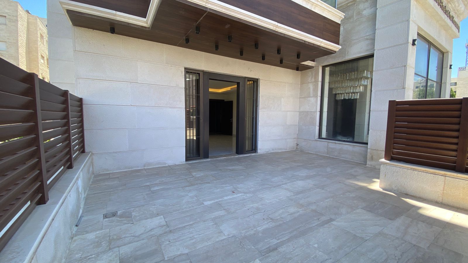 260ca014-c02f-4ce0-b6cd-2bf12ca0c47a Outdoor tiled patio with beige stone walls, brown privacy fencing, and double glass doors opening to an interior space.