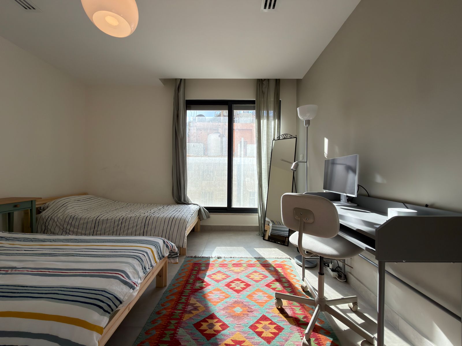 26206b67-439e-4e3f-a206-772a8850abce Small bedroom with two single beds, striped bedding, and a colorful geometric rug under the window. A desk with monitor and a swivel chair sits to the right, plus a standing mirror and lamp by the window.