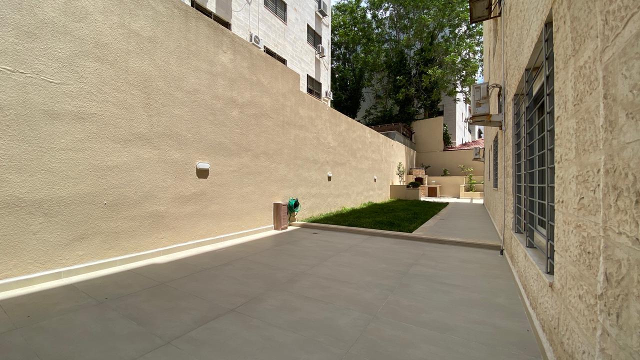 2746cb0c-d3c3-44fe-9151-4f85f663b0c5 Sunny back courtyard between beige buildings with a grey tiled patio, a small grassy strip, and barred windows.