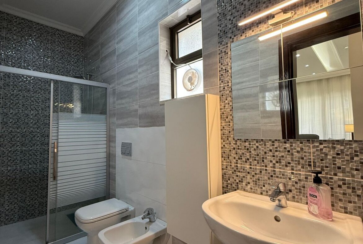 Modern bathroom with glass shower enclosure, mosaic tile walls, and a white pedestal sink with pink hand soap.