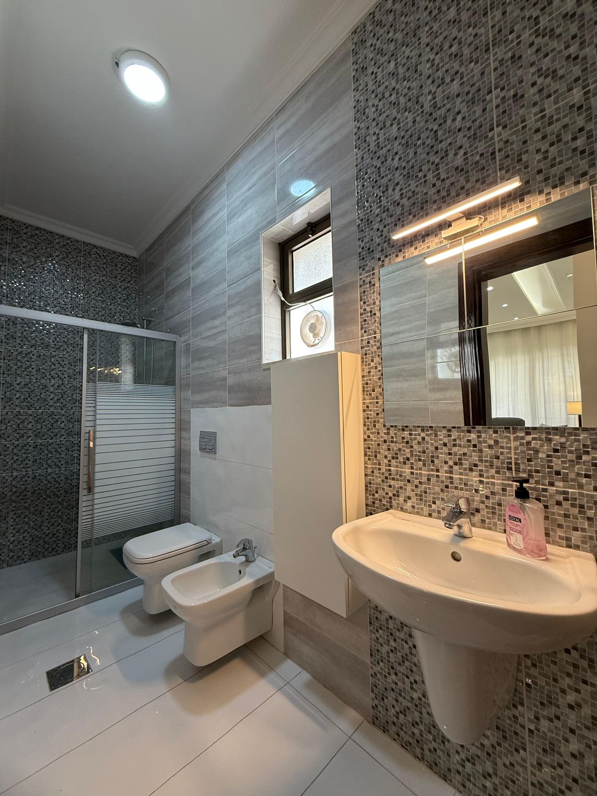 Modern bathroom with glass shower enclosure, mosaic tile walls, and a white pedestal sink with pink hand soap.