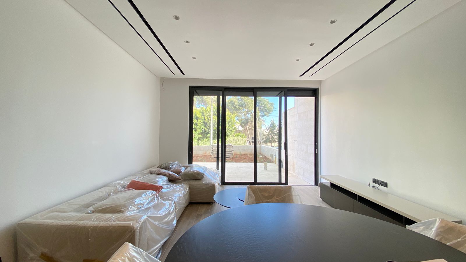 274d597e-7def-4d2a-b2bc-24dfc5275b82 Modern white living room with plastic-covered sofas and a dark round table, sliding glass doors to a patio and garden beyond.