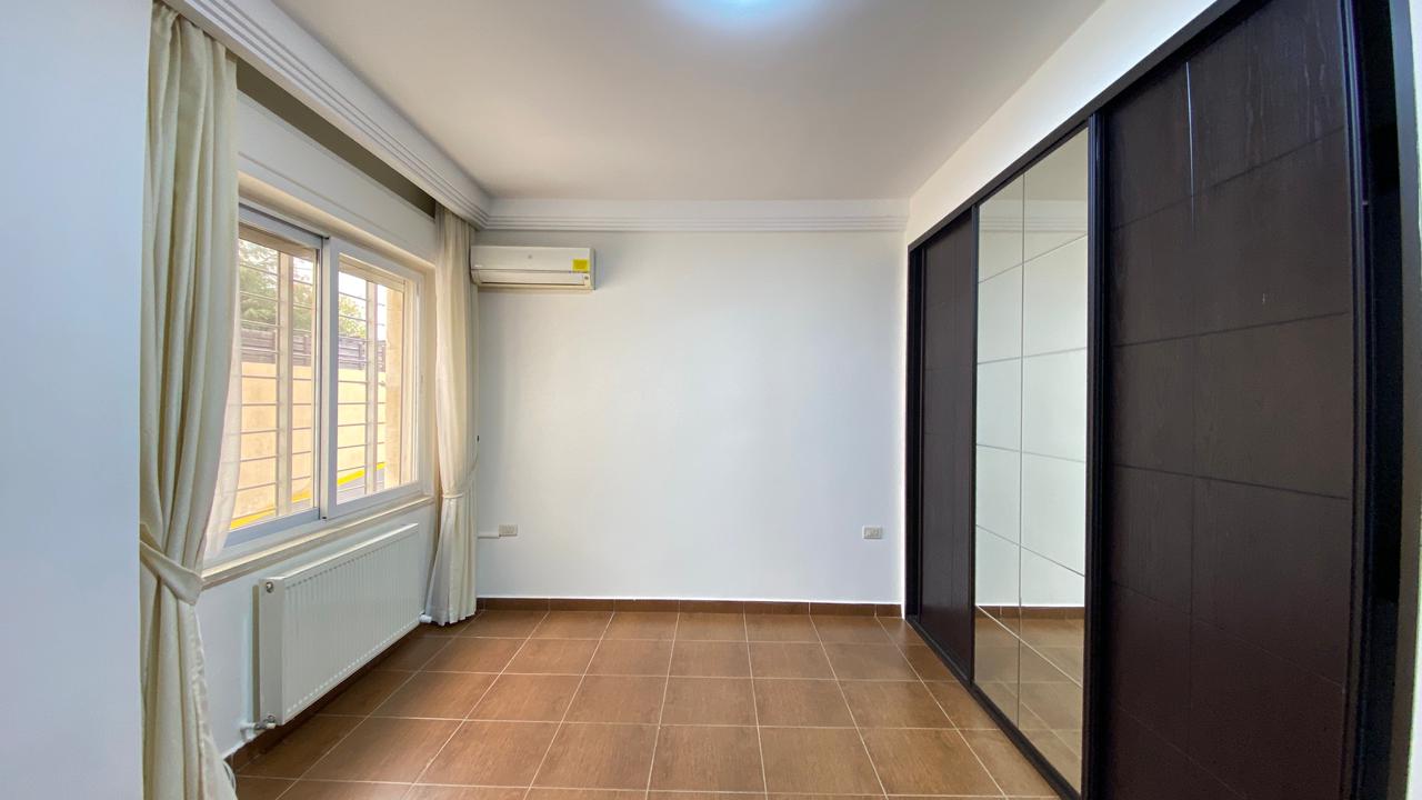 278c43bd-cd8a-441e-8649-b13560697c75 Bright empty bedroom with brown tiled floor, a large window with cream curtains, and a built-in wardrobe with sliding mirrored doors on the right.