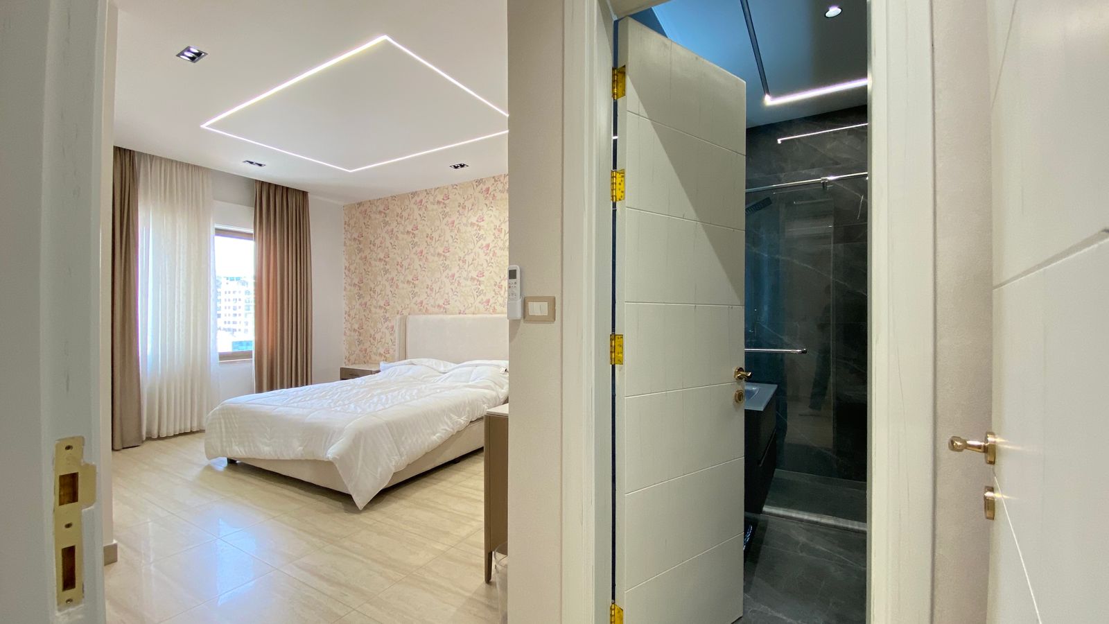 27be4912-261c-4432-a8ff-fce256b0f144 Modern bedroom with white bed and floral accent wall; large window with beige curtains and geometric ceiling lights visible from doorway to bathroom
