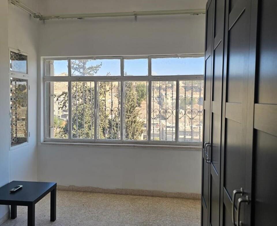 280b465e-7ce4-4c2f-8de5-d66b4265a842 Bright bedroom with a large window, view of trees and hills, and a dark wardrobe along the right wall; a small black table and a bed are visible.