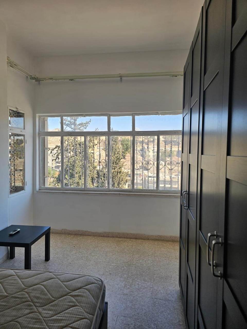 280b465e-7ce4-4c2f-8de5-d66b4265a842 Bright bedroom with a large window, view of trees and hills, and a dark wardrobe along the right wall; a small black table and a bed are visible.