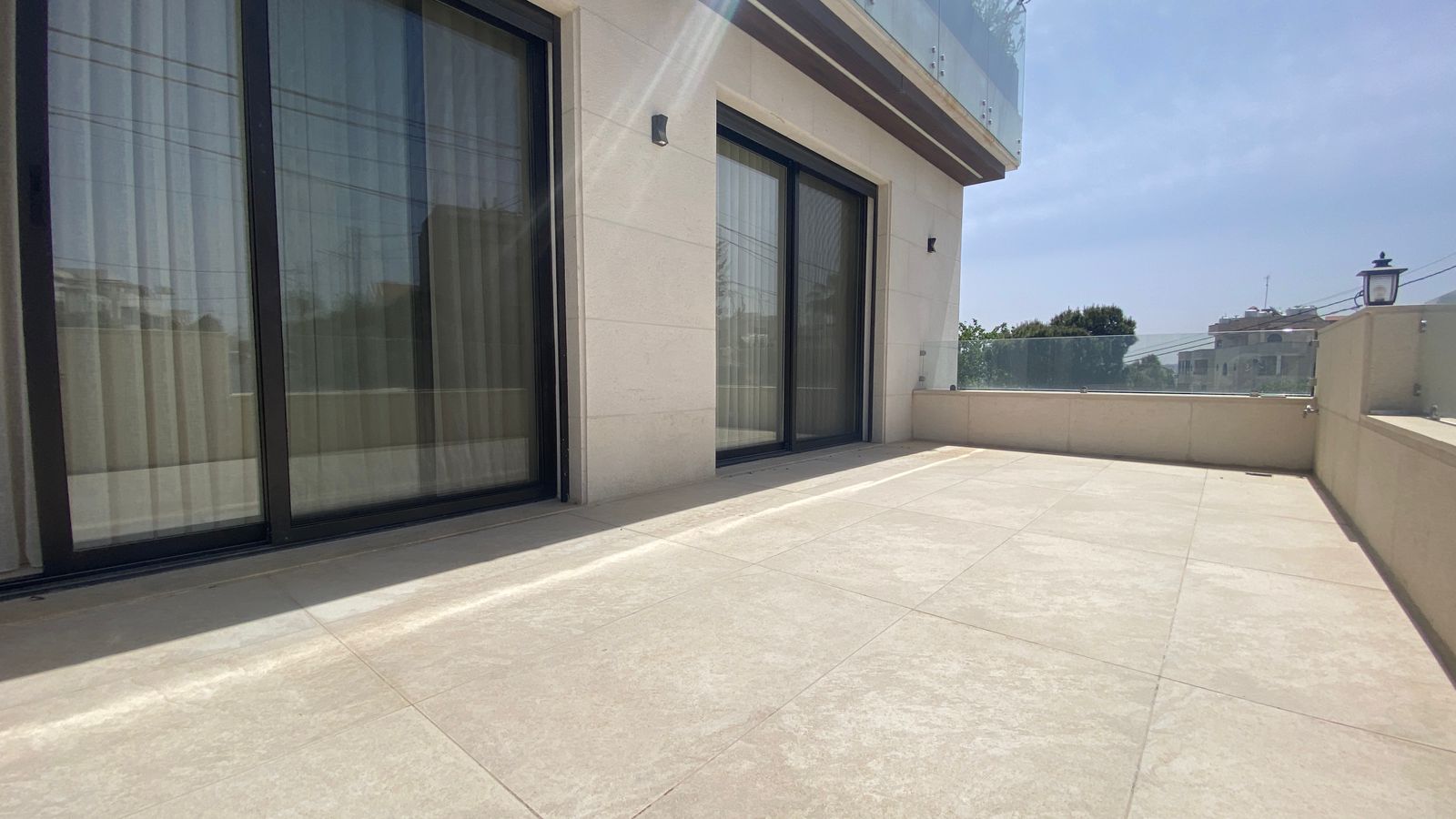 280f9ace-4271-401c-938d-5c756cbc4b39 Sunlit rooftop terrace with beige stone walls, large sliding glass doors, and a glass railing overlooking the neighborhood.