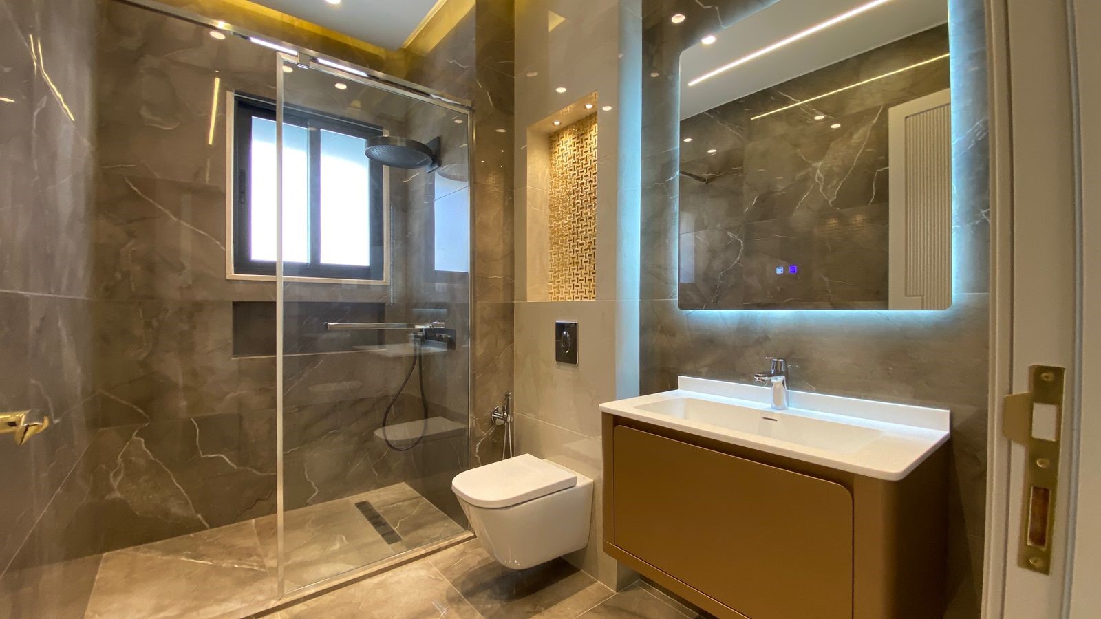28304d09-b2ad-4c2e-896c-b87849a6edc3 Modern bathroom with a glass-enclosed shower, wall-hung toilet, and brown vanity under a backlit mirror in marble-tiled surroundings.