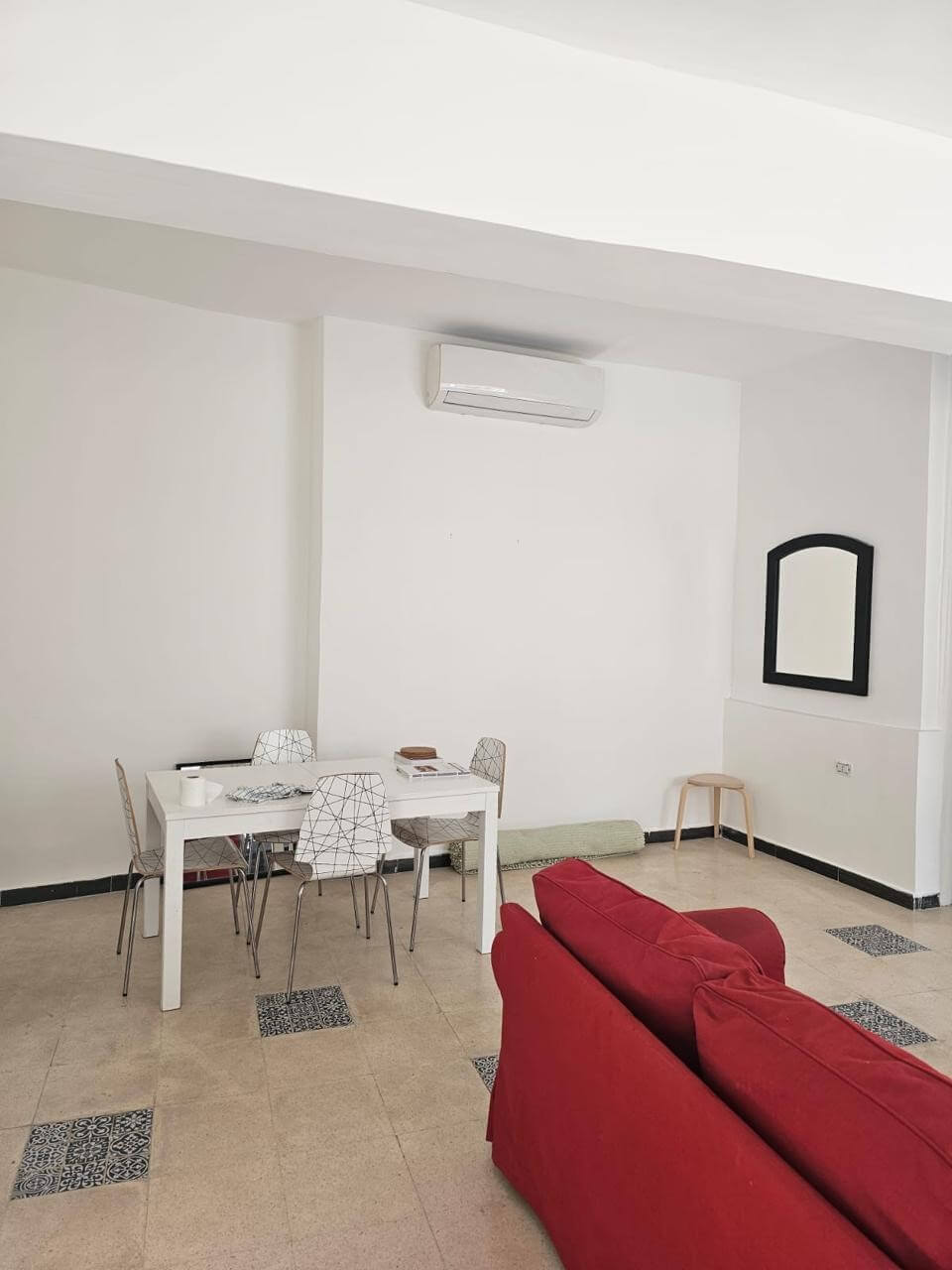 2833e65e-fd54-4b63-a7b2-7a7a69843f18 White minimalist dining area: a white table with four wireframe chairs, a red sofa in the foreground, and a wall mirror with a black frame.