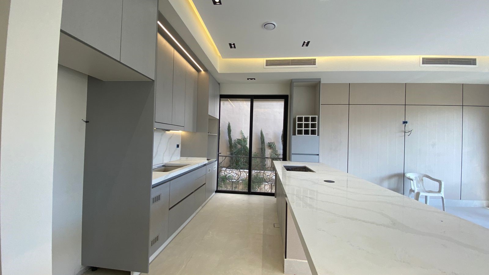 28702093-442c-402d-bd4f-ed6bc412e431 Contemporary kitchen with light gray cabinetry, marble countertops, and a large island with a built-in cooktop, near a glass door leading to an outdoor courtyard.