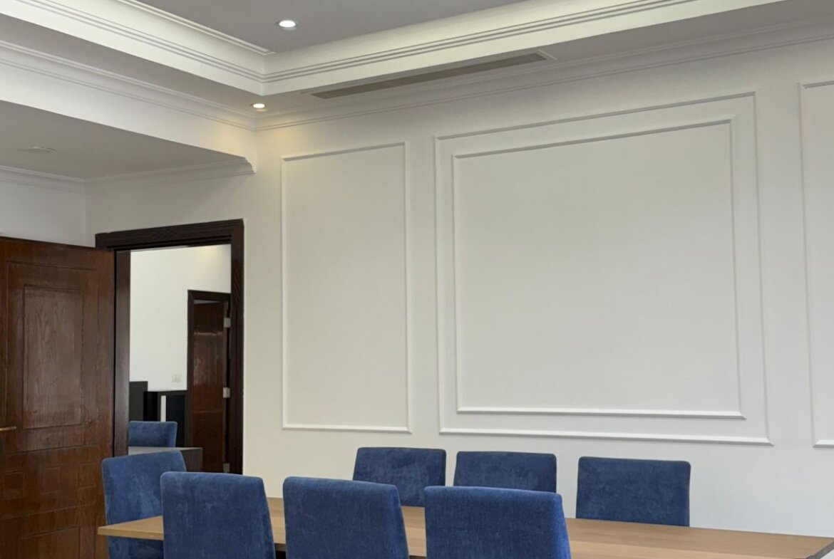 A conference room with a wooden table and blue upholstered chairs along white paneled walls and a wooden door in the background.