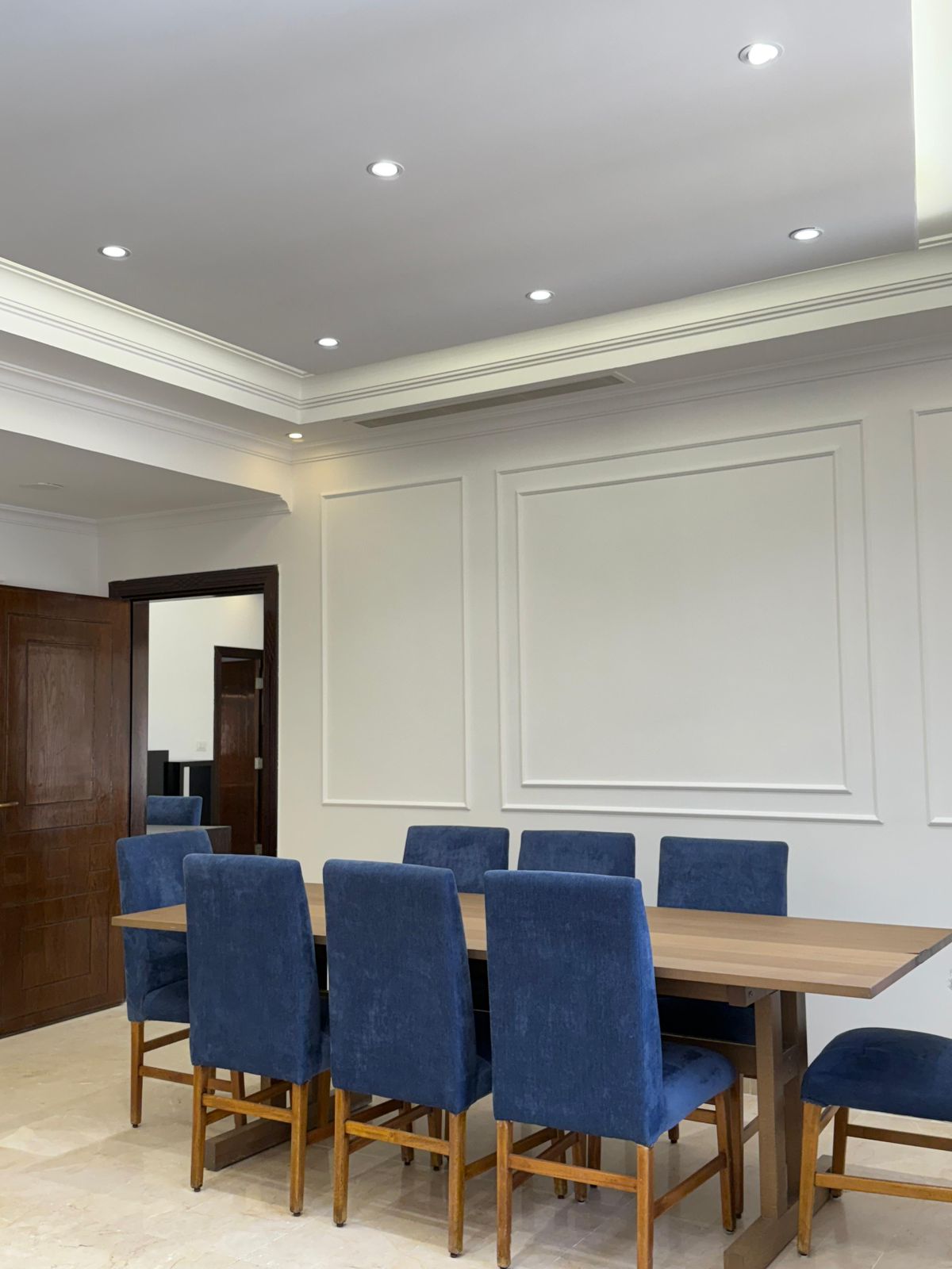 A conference room with a wooden table and blue upholstered chairs along white paneled walls and a wooden door in the background.