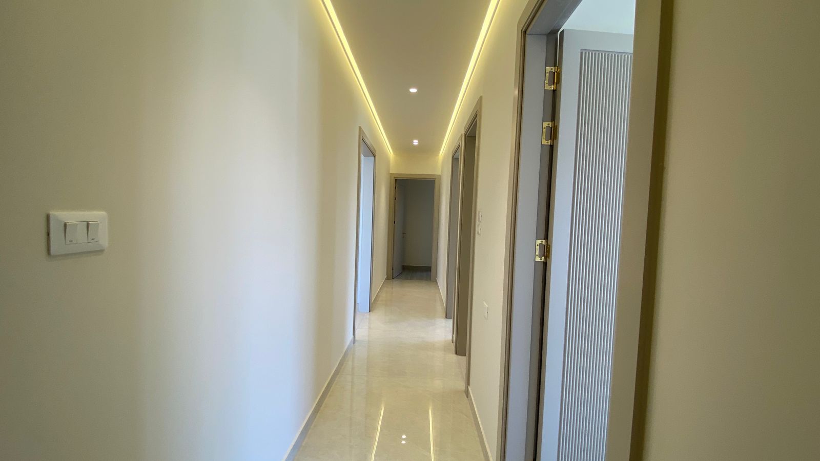 29791598-5687-4295-a146-80ae3e2a3016 Long, well-lit hallway with cream walls, a glossy tiled floor, and doors along both sides leading to rooms.