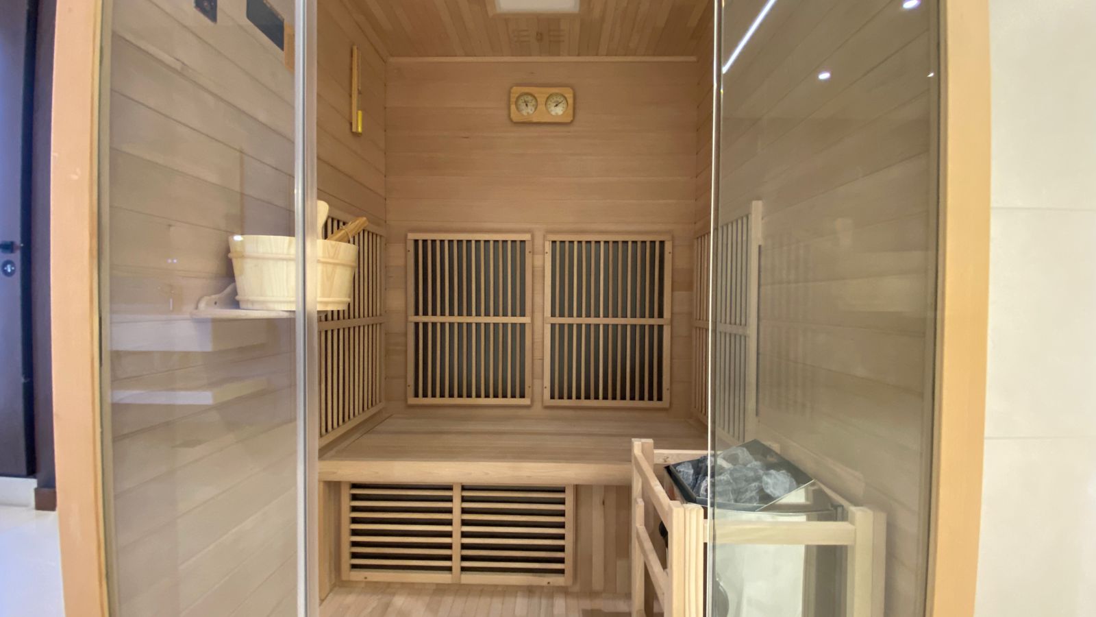 297a5843-9a5e-486f-8eb2-7fd04dd967f4 Inside a wooden sauna: bench seating, a heater with stones, and a bucket on the shelf behind glass doors