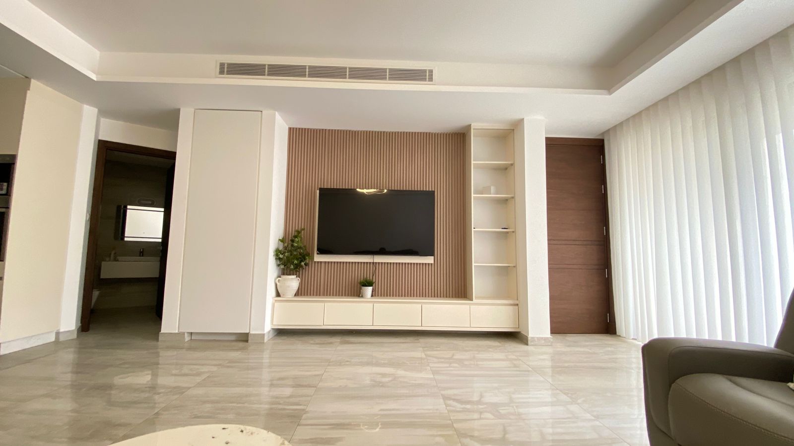 29b4f4ae-5f49-4d2d-83ff-f8dfadae8172 Modern living room with a mounted flat-screen TV on a wood-textured backdrop, white media console and open shelves across a minimalist entertainment wall.
