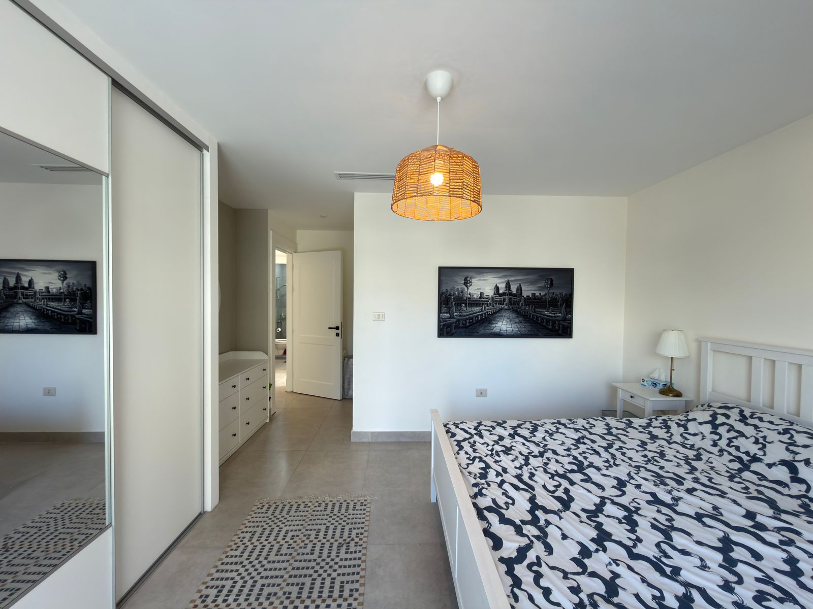 29ea4932-d5b0-43ba-a090-38118c88c9d5 Bright bedroom with white bed frame, blue‑white patterned duvet, wicker pendant light, and black‑and‑white framed landscape art on the wall