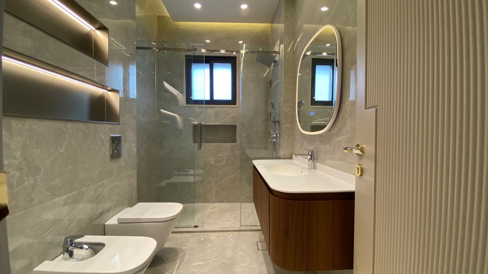 2a5db346-12d1-4928-bde1-6ebbfaeb388d Modern bathroom with glass shower enclosure, wall-mounted toilet and bidet, and a curved wood vanity with a white sink.