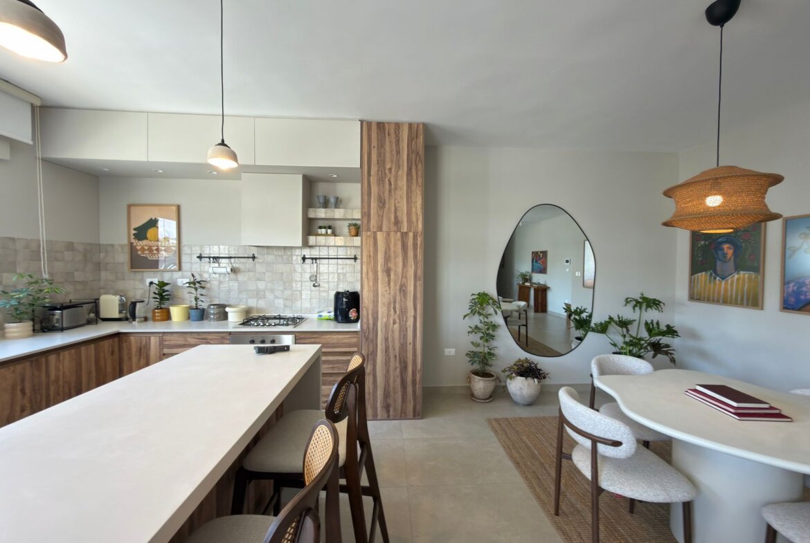 Open kitchen and dining area featuring a white island, wood cabinets, beige tile backsplash, and pendant lights; a round mirror and dining table are nearby with plants in the scene.