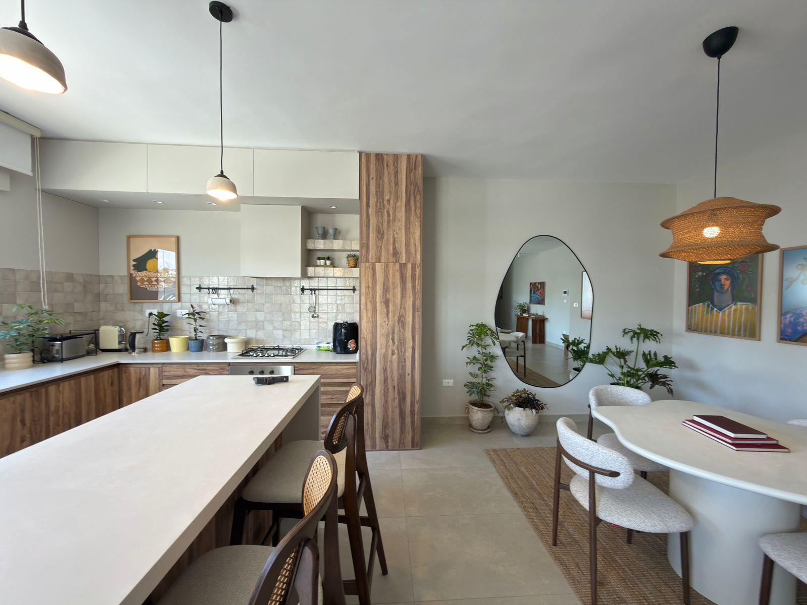 2ae23eb4-a10a-4179-9272-79c6387d8c39 Open kitchen and dining area featuring a white island, wood cabinets, beige tile backsplash, and pendant lights; a round mirror and dining table are nearby with plants in the scene.
