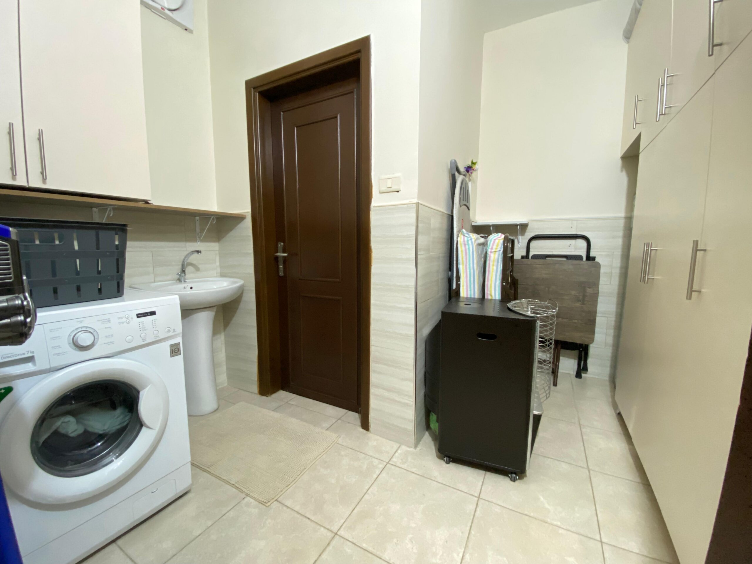2af401d1-9a78-4e27-8c5b-3a8a43a52c74 Laundry room with washer, sink, and white cabinets, a brown interior door, and a rolling laundry cart nearby.