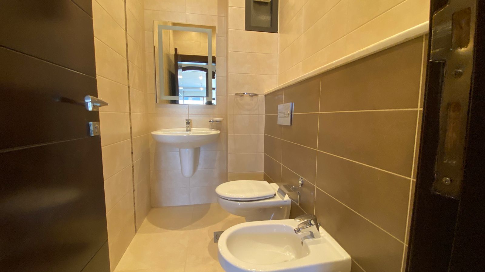 2b03206a-90d3-4954-b317-0ee71ede2eb1 Small bathroom with beige wall tiles, a pedestal sink, a wall-mounted mirror, a toilet, and a bidet visible near the foreground.