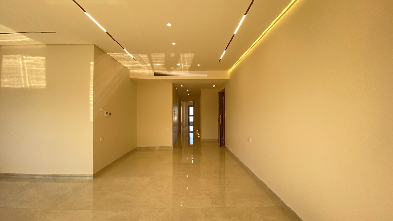 2b0c0ed7-25bc-49be-b1b2-45f56c83c0c1 Long empty corridor in a modern building with glossy tiled floor and warm yellow lighting, beige walls, and ceiling strip lights.