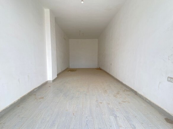 Long empty interior room with white walls, pale tiled floor, and a single exposed light bulb hanging from the ceiling at center. It looks unfinished and clean but dusty.
