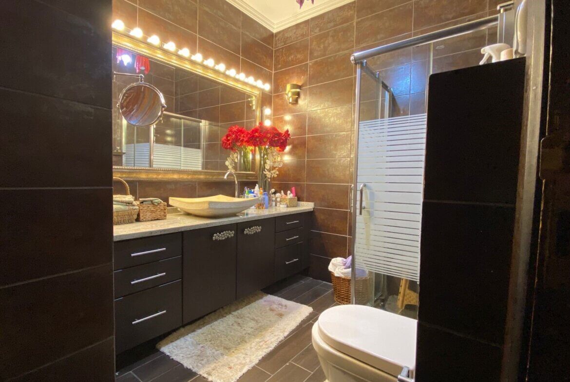 2c4d6e6c-d9c5-435e-89f7-42224adf3eb7 Dark tiled bathroom with a wide vanity, vessel sink, and glowing mirror lights; red flowers centerpiece on the counter as a shower and glass door appear to the right.