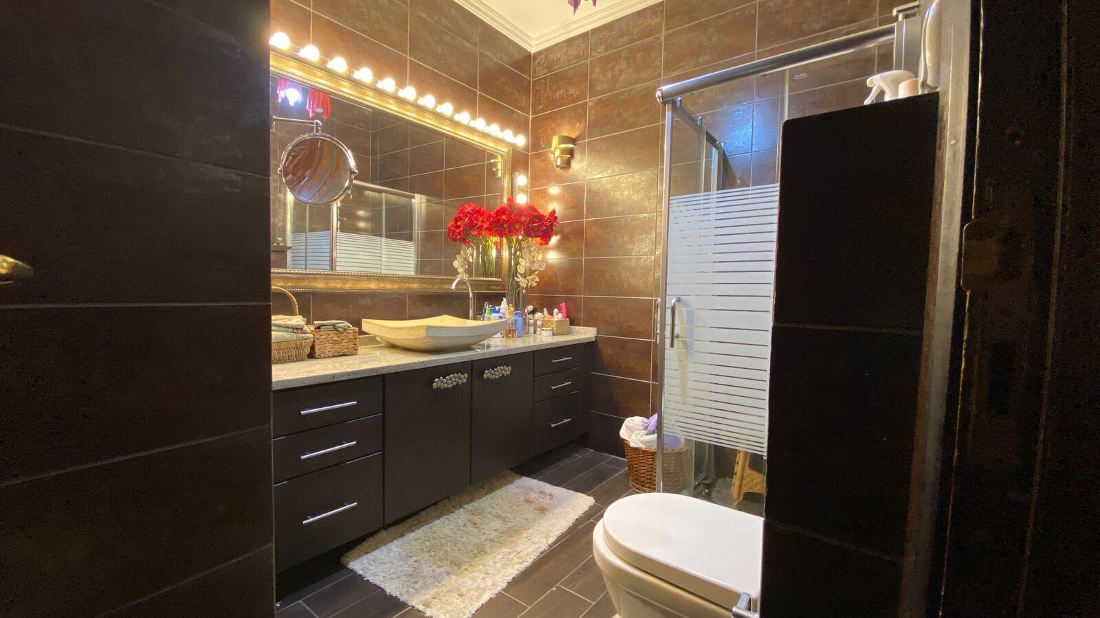 2c4d6e6c-d9c5-435e-89f7-42224adf3eb7 Dark tiled bathroom with a wide vanity, vessel sink, and glowing mirror lights; red flowers centerpiece on the counter as a shower and glass door appear to the right.