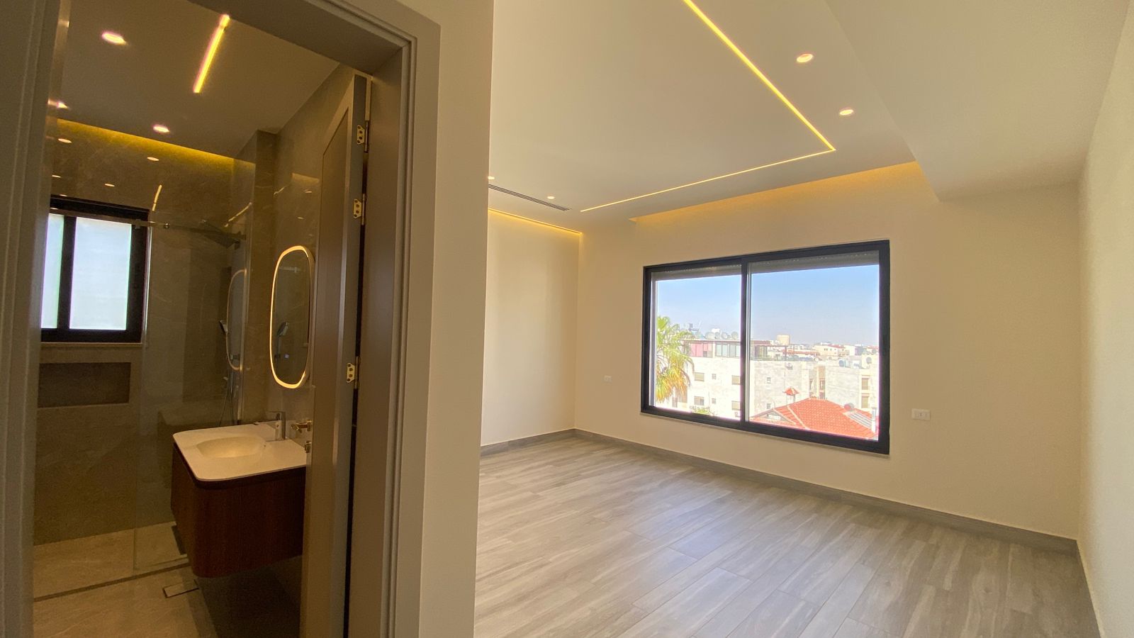2ce9f3c9-f5cb-4546-8785-a4fe7f7ebb7e Bright empty room with a large window showing a city view, light gray wood floor, and recessed ceiling lighting; bathroom doorway on the left.