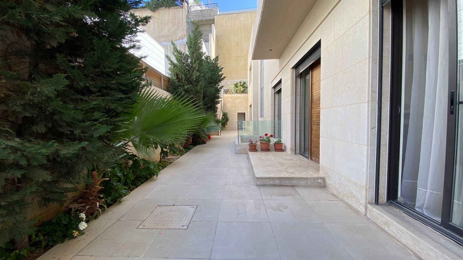 2d624178-9016-4460-b9a7-5b9381f24bd3 Narrow outdoor walkway beside a beige stone building, with potted flowers and tall evergreens along the left wall.