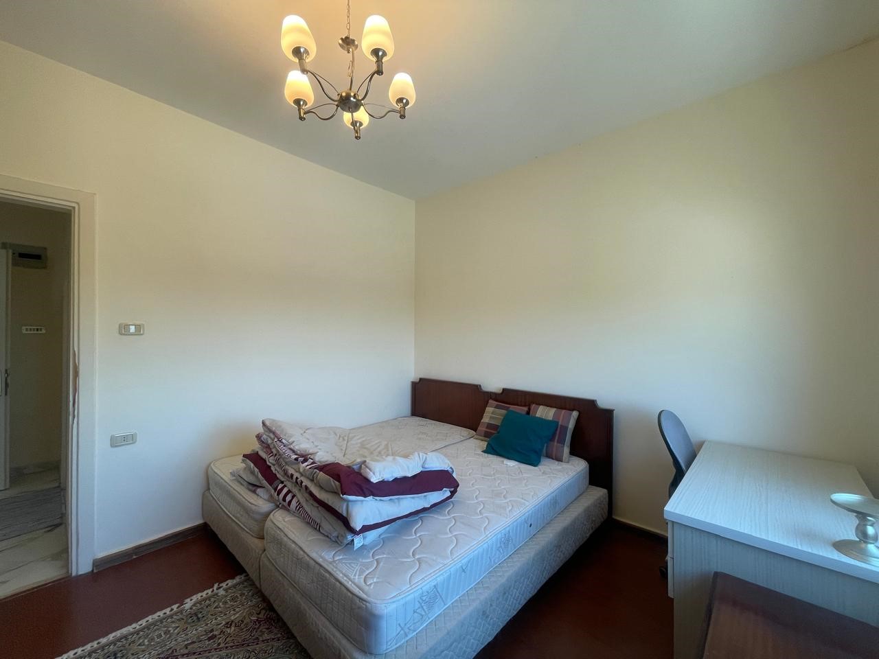 2f5448b5-f3a4-4d92-aa6f-1d4c32ac9e86 Bedroom with a bed against the wall, stacked blankets and pillows, and a white desk with a chair to the right under a chandelier.
