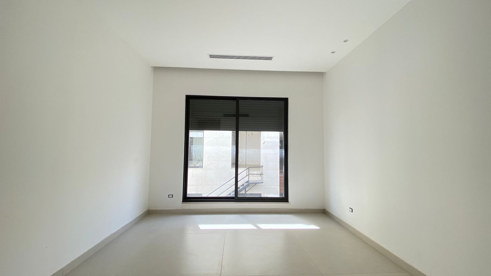 2fab1206-5208-4758-93b0-6f1dd067f1f4 Empty white rectangular room with a large black-framed window centered on the far wall, sunlight casting a strip of light on the tiled floor outside view shows a metal staircase railing .