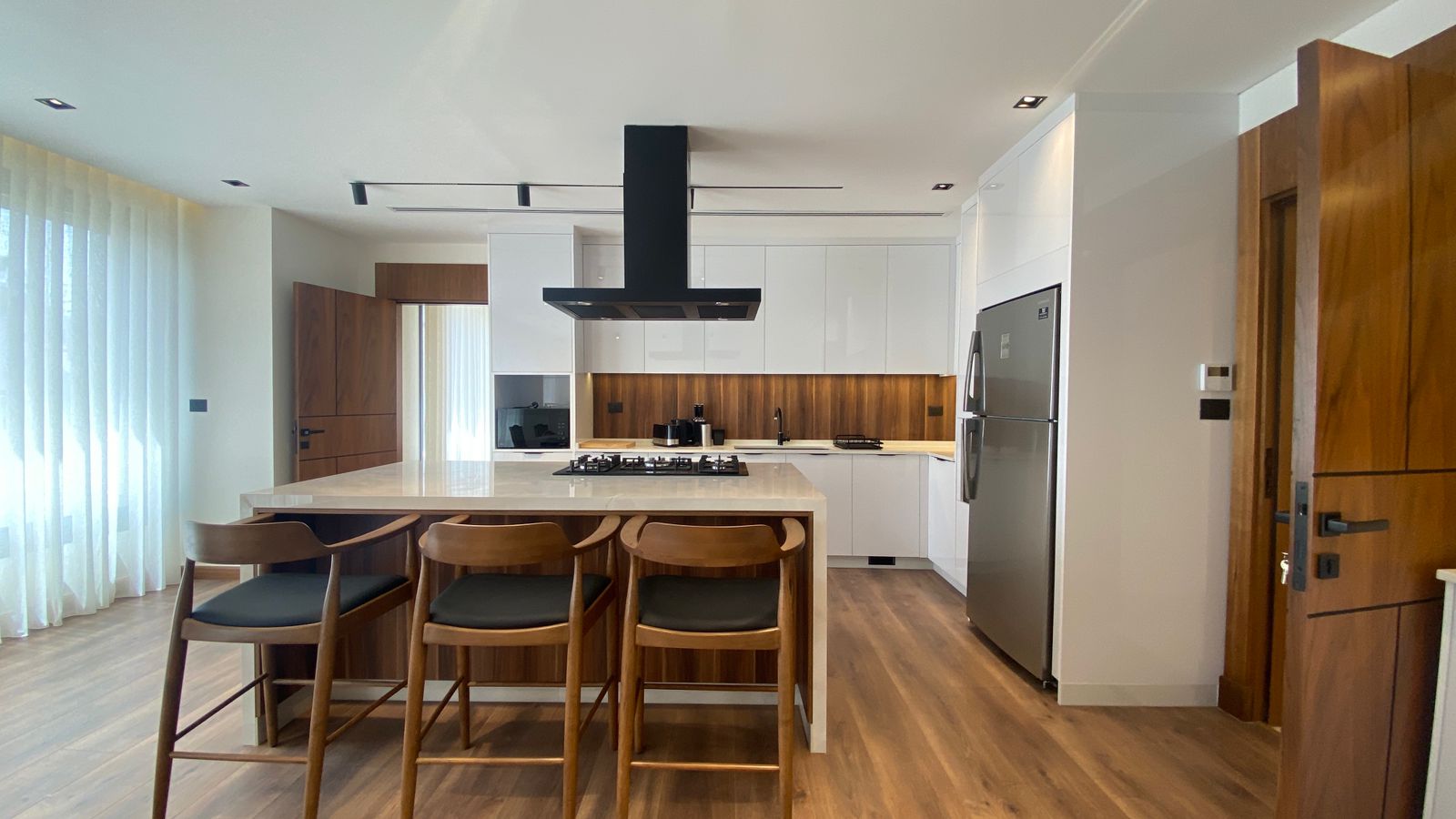 30326cb3-4929-49fe-a989-ce8a45a788ef Modern kitchen with white cabinetry, wood backsplash, and a central island with four wooden stools.