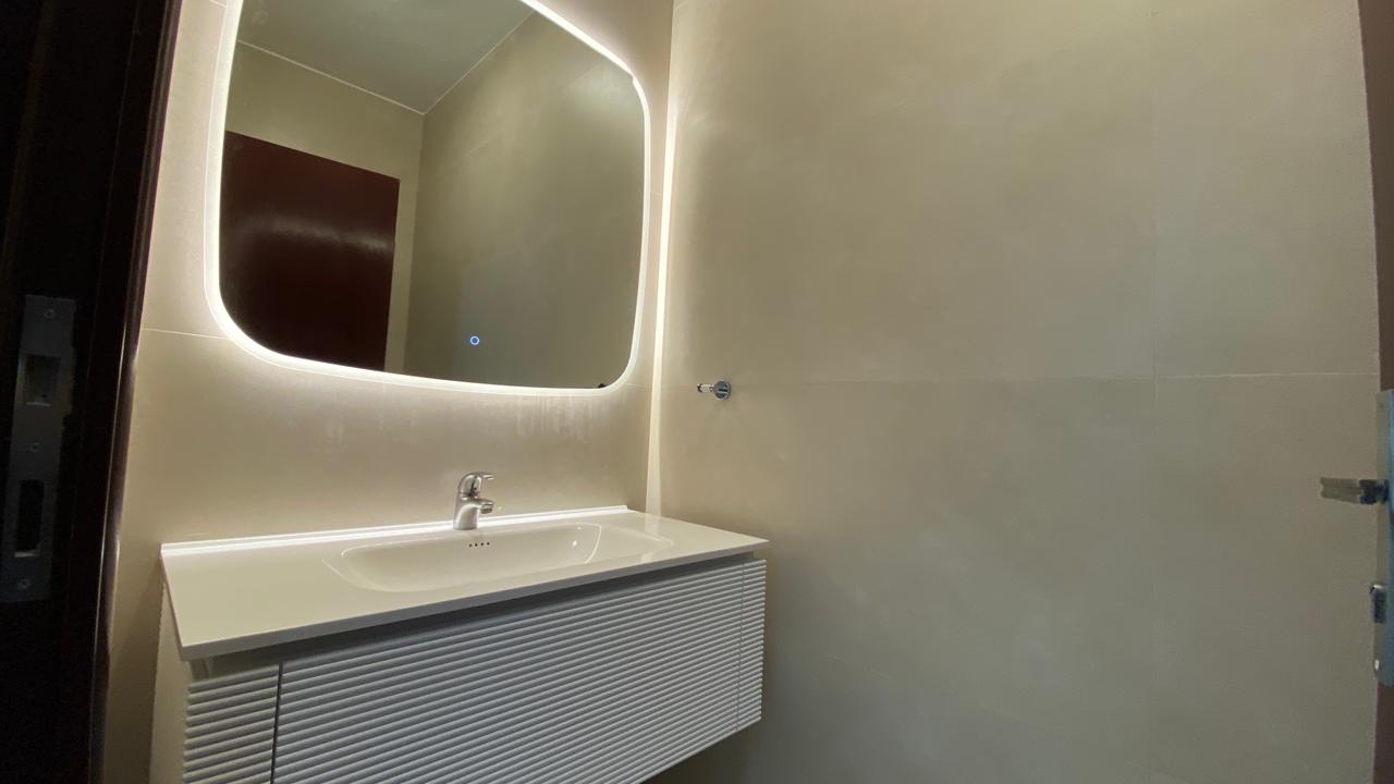 30b641c7-6ffe-42d0-b88e-d7ff3cb85aa3 Modern bathroom vanity with a backlit rounded rectangular mirror, white sink, and chrome faucet.