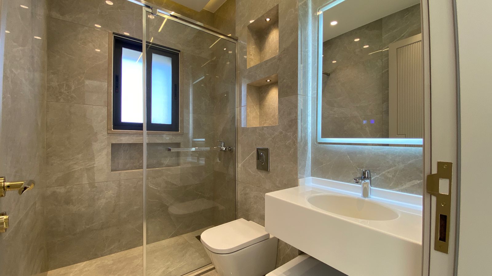312ea571-a91c-4ed5-8861-99711413614a Modern marble-tiled bathroom with a glass shower enclosure, wall-mounted toilet, and white rectangular sink under a backlit mirror.