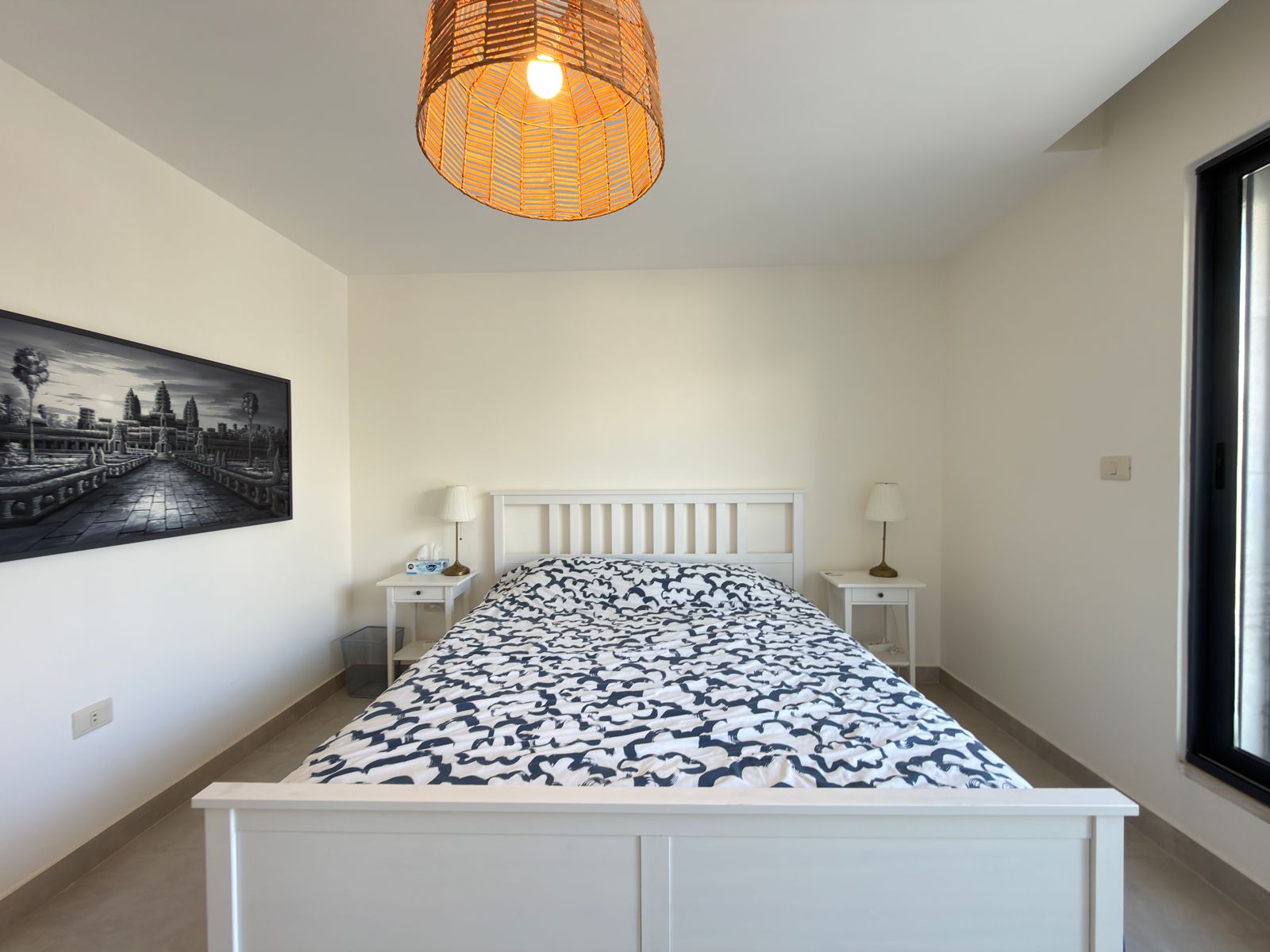 316dffba-836f-4585-a490-f4316f7aa98b White wooden bed with a blue-and-white patterned duvet, flanked by two white nightstands and matching lamps in a bright minimalist bedroom.