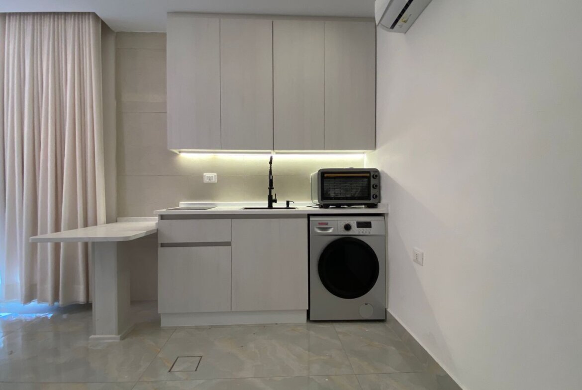 Modern gray kitchen with a fold-out counter, sink with black faucet, under-cabinet lighting, and a washing machine beside a toaster oven on the counter.