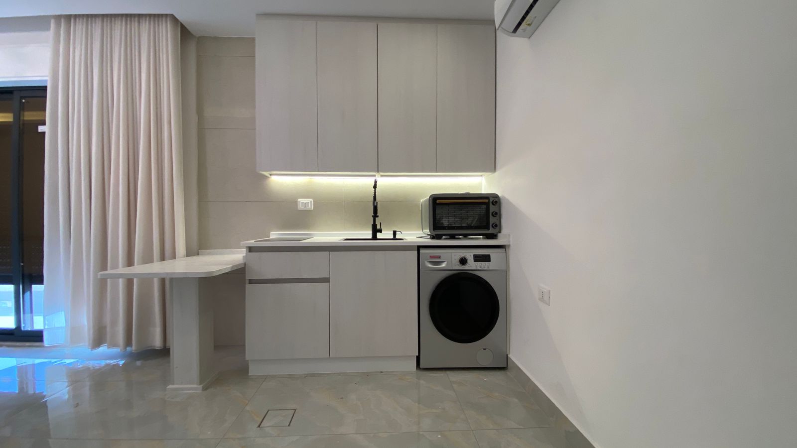 Modern gray kitchen with a fold-out counter, sink with black faucet, under-cabinet lighting, and a washing machine beside a toaster oven on the counter.