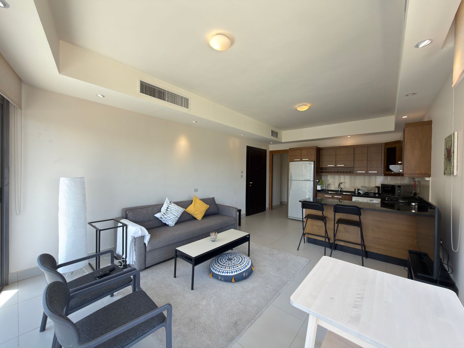 32e97e1c-3a55-46f0-b5cd-73e0065562a8 Open-concept living area with gray sofa, black coffee table, and a round patterned pouf facing a kitchen island and bar stools.