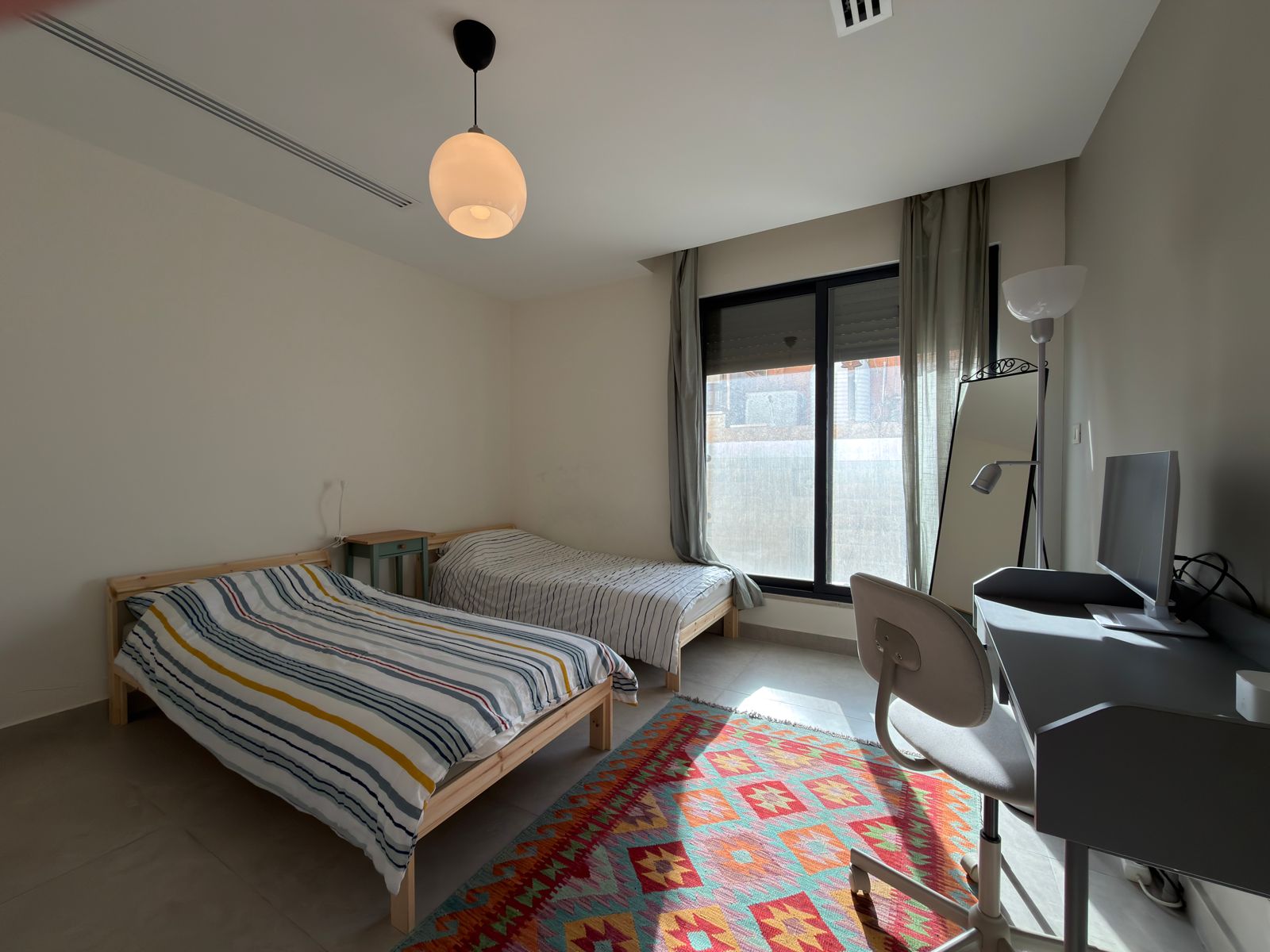331aa27c-3b4a-4ea1-9a20-9baba59d4b2d Light-filled bedroom with two single beds, striped bedding, and a colorful geometric rug; a window and a small study area are visible by the desk and chair.