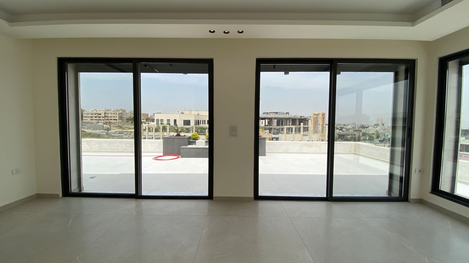 33b1b189-79ec-44fb-aea0-8857cb19c71b Empty living room with three large sliding glass doors opening to a sunlit terrace and cityscape beyond