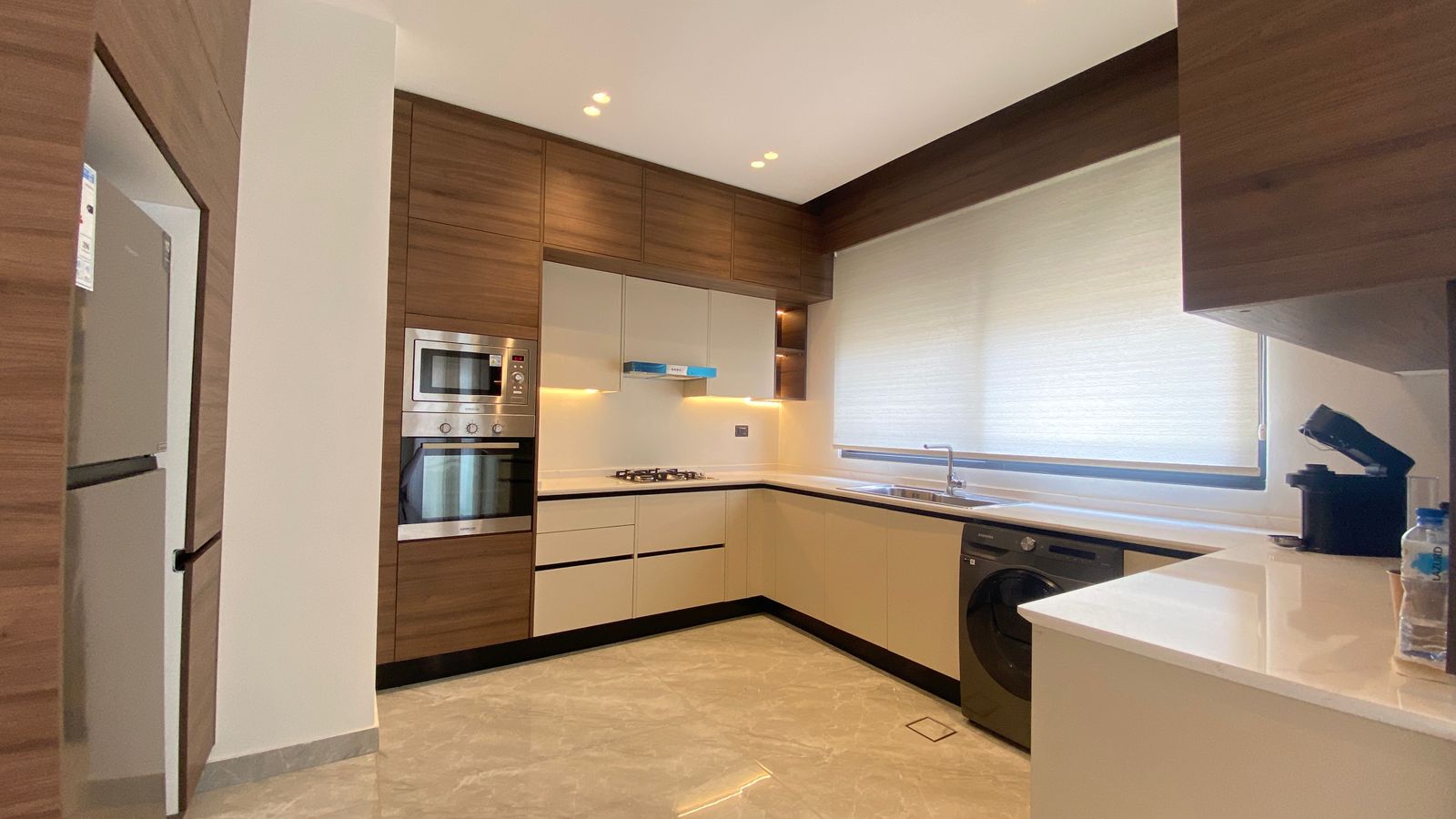 33e6040a-0e04-4f5b-b4d5-04678809935e Modern L-shaped kitchen with wood cabinets, white countertops, built-in stainless steel oven and microwave, gas cooktop, and washing machine nearby.