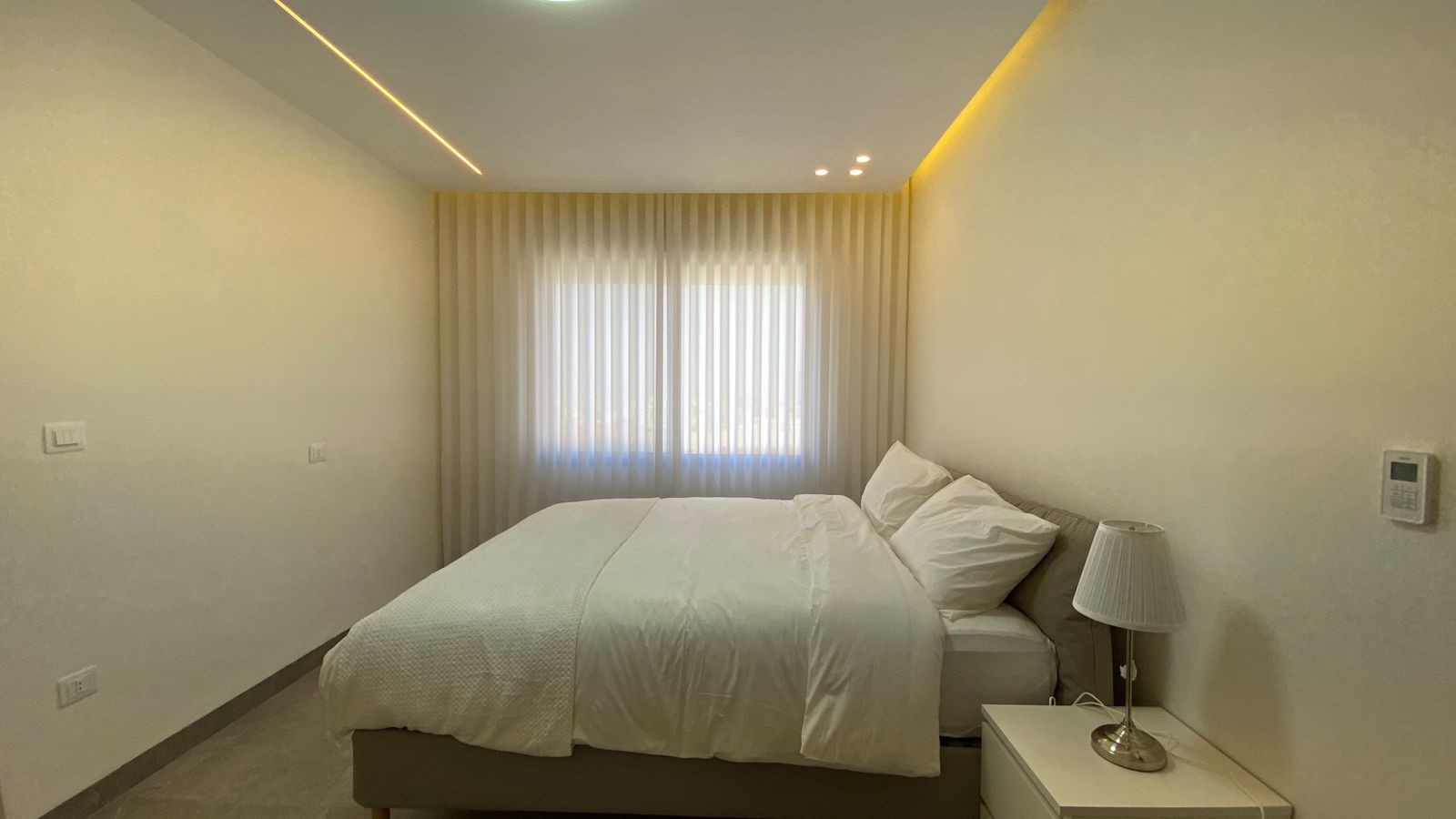 34581d54-9d5a-4173-bbb0-a889b3f23ea2 Minimalist hotel bedroom with a large bed, white bedding, and a nightstand with a lamp beside a window with sheer curtains