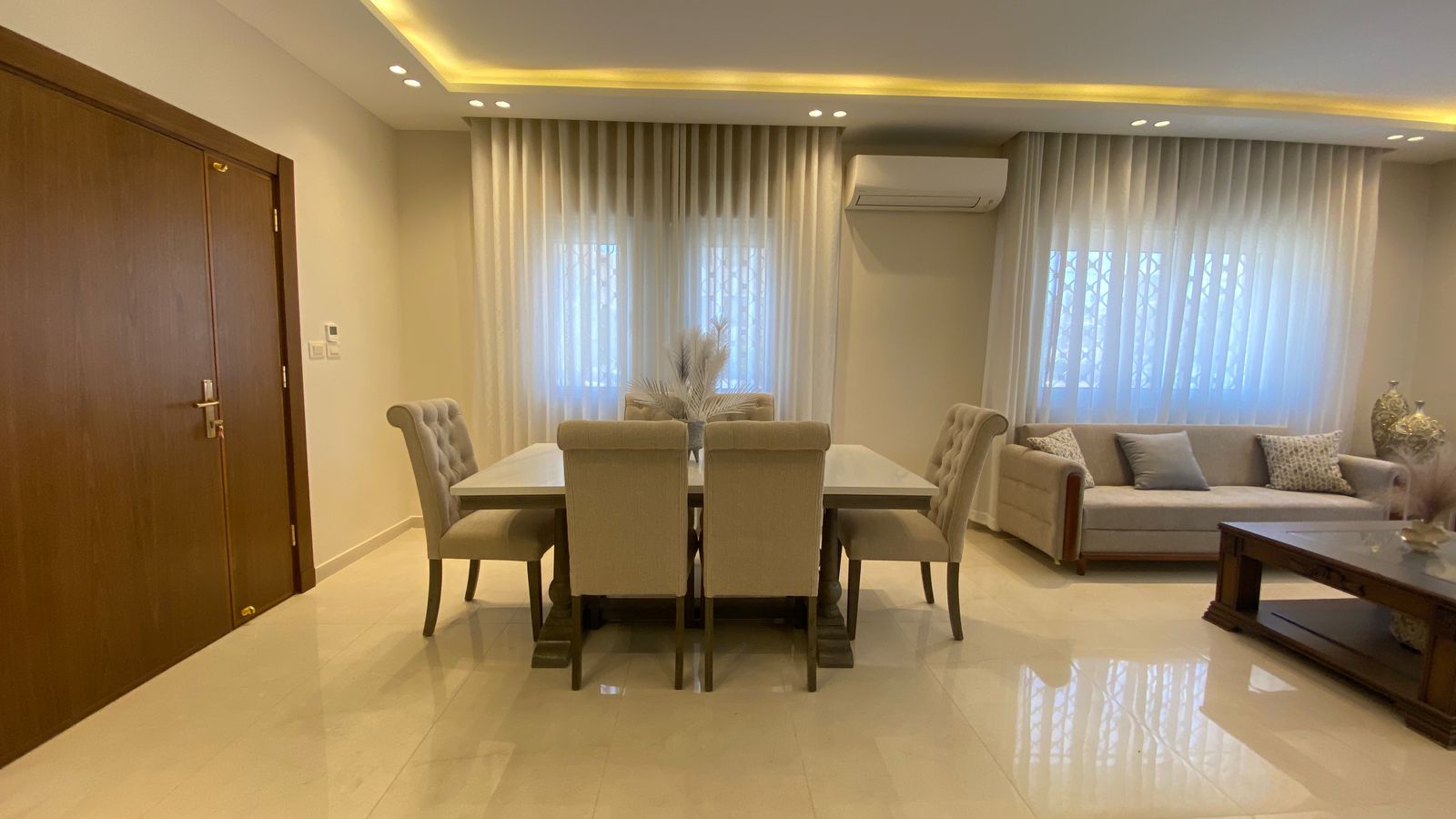 347c6cf3-3fff-4e5e-ac5b-7766c003004d Open-plan living and dining room featuring a white dining table with six beige upholstered chairs, a beige sofa with cushions, and sheer curtains by large windows.