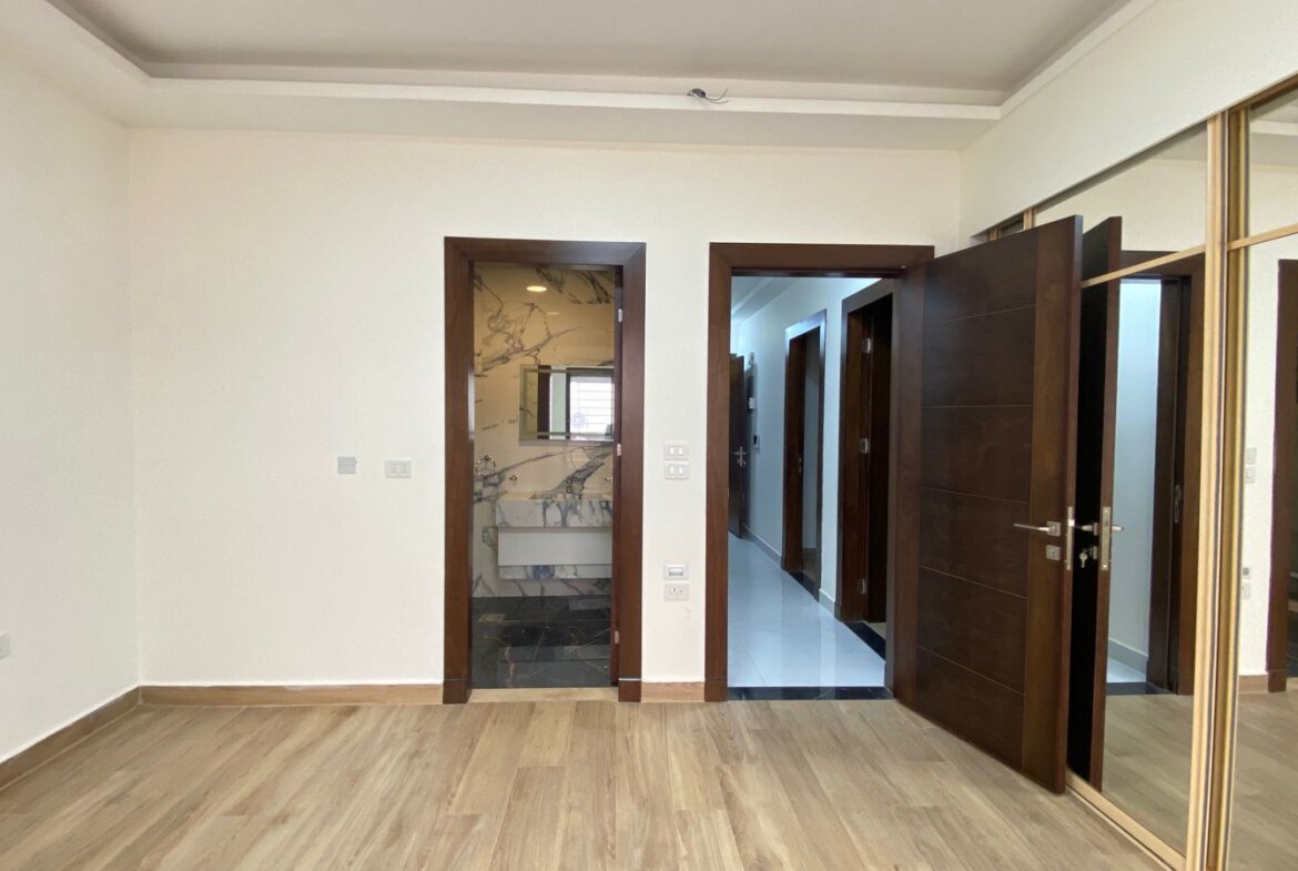 Empty room with light wood flooring and three doorways: one to a marble-floored bathroom, one to a hallway, and a large mirrored wardrobe along the right wall.